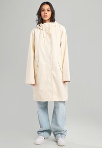 Cream-colored waterproof coat with a high collar, front buttons, two side pockets, and long sleeves, paired with light blue wide-leg jeans.