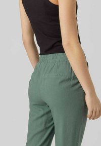 Groene, elastische taillebroek met een gladde textuur, achterzakken en een relaxte pasvorm, gecombineerd met een aansluitend zwart tanktop.