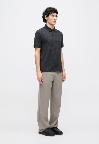 Vīriešu modelis valkā melnu polos krekliņu ar rakstu un īsām piedurknēm, kopā ar gaiši pelēkām biksēm un melnām sporta kurpēm.