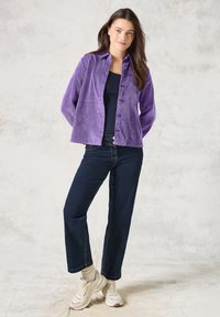 Giacca a bottoni in corduroy viola abbinata a jeans in denim scuro a taglio dritto, indossata con sneakers chiare e una maglietta blu navy sotto.