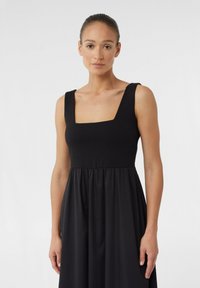 Schwarzes ärmelloses Kleid mit quadratischem Ausschnitt, tailliertem Oberteil und fließendem Rock. Hergestellt aus weichem Stoff mit glatter Textur.