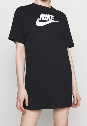 Persoon draagt een losse zwarte Nike T-shirt jurk met wit logo, staand tegen een effen lichte achtergrond, armen ontspannen langs het lichaam.