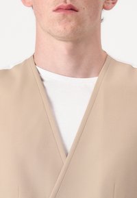 Gilet avvolgente beige con scollatura a V, indossato su una maglietta bianca semplice, caratterizzato da un tessuto morbido e dettagli di cucitura delicati.