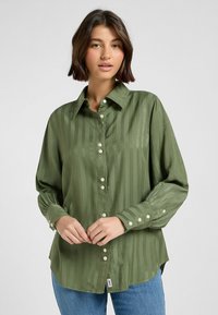 Camicia button-up verde in satin con righe verticali, maniche lunghe e colletto classico. Presenta bottoni bianchi e un orlo leggermente asimmetrico.