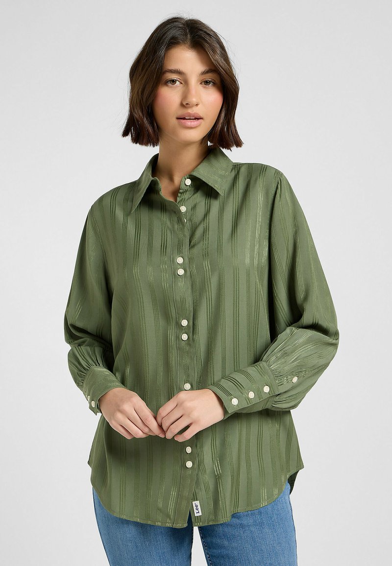 Camicia button-up verde in satin con righe verticali, maniche lunghe e colletto classico. Presenta bottoni bianchi e un orlo leggermente asimmetrico.