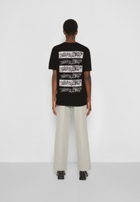 Osoba stoi tyłem, ubrana w czarną koszulkę z powtarzającym się białym nadrukiem oraz jasnobeżowe szerokie spodnie i czarne buty.