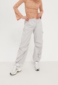 Světle šedé cargo kalhoty s elastickým pasem, zúženými nohavicemi a bočními kapsami, v kombinaci se stříbrnými sportovními botami a texturovaným oranžovým topem.