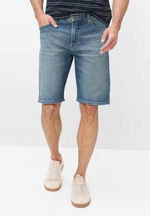 Mann trägt mittellange blaue Jeans-Shorts, schwarzes gestreiftes Shirt und weiße Sneakers, steht mit einer Hand in der Tasche.