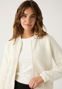 Eine Frau mit welligem braunem Haar trägt eine weiße gesteppte Zip-Jacke über einem weißen Oberteil und posiert vor einem schlichten hellen Hintergrund.