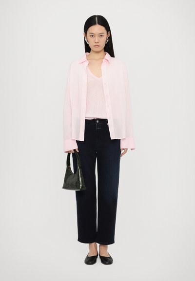 Chemise boutonnée rose clair sur un top rose clair, jean droit en denim foncé, ballerines noires et un sac à main en croco embossé vert foncé.