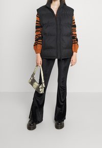 Frau in schwarzer gepolsterter Weste und schwarzen samtigen Schlaghosen, die eine kleine Handtasche mit Schlangenprint vor einem einfarbigen grauen Hintergrund hält.