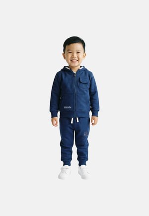 Jeune garçon souriant, portant un sweat à capuche zippé bleu marine et un pantalon de survêtement assorti, debout avec des baskets blanches sur un fond uni.