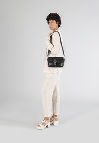 Svart läder axelväska med klaffstängning och metalldetaljer, i kombination med en ljus, texturerad, off-white outfit och vita klacksandaler.