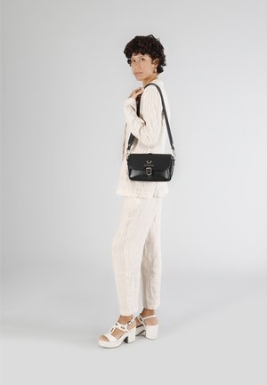 Sort læder skuldertaske med klaplukning og metalhardware, parret med et let, struktureret, off-white outfit og hvide hælsandaler.