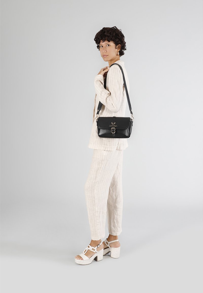 Svart läder axelväska med klaffstängning och metalldetaljer, i kombination med en ljus, texturerad, off-white outfit och vita klacksandaler.