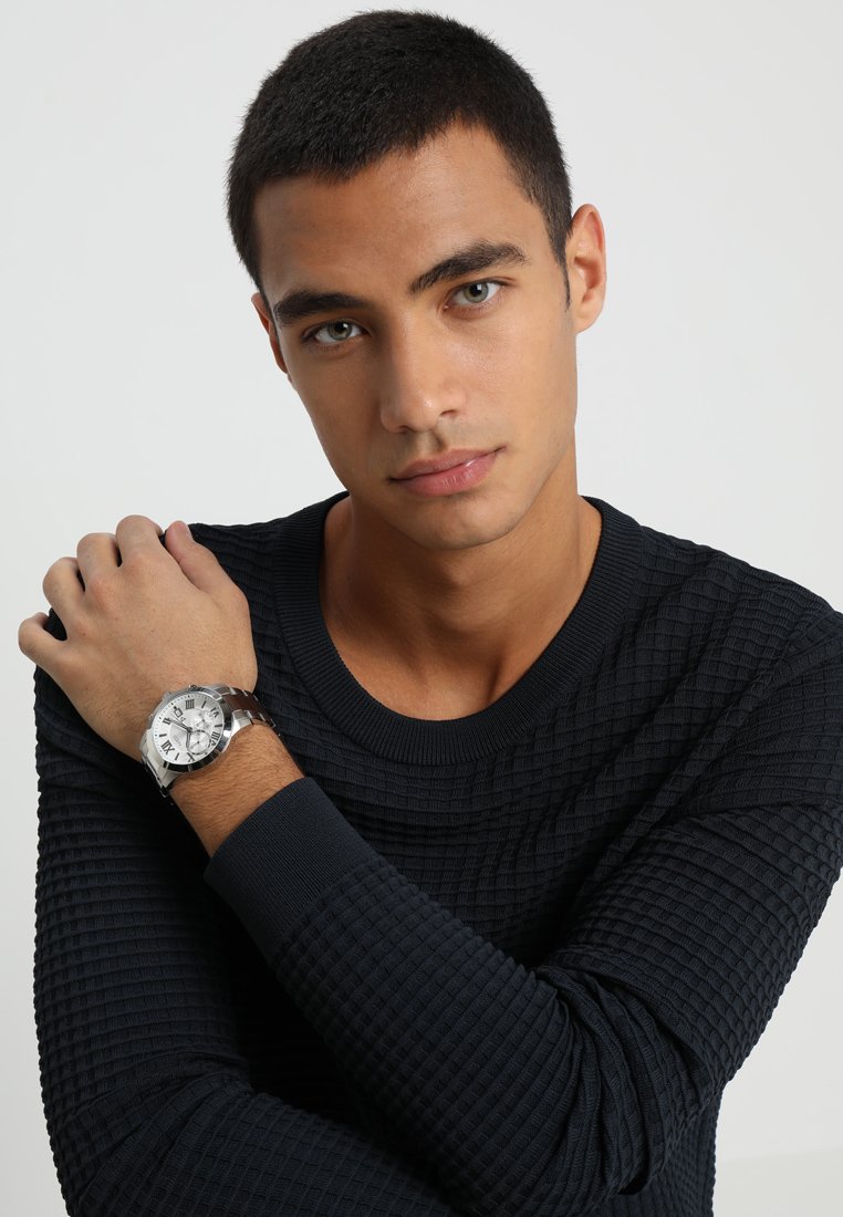Montre en argent avec un cadran rond, des chiffres romains et un bracelet en mailles. Le modèle porte un pull sombre texturé avec un col rond.
