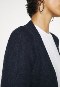 Granatowy sweter na drutach z miękkim, strukturalnym wykończeniem, otwarty z przodu, ze ściągaczami na mankietach, noszony na białej koszuli. Widoczna duża srebrna kolczyk koło.