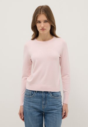 Jeune femme aux cheveux lisses châtain clair, portant un pull à manches longues rose clair et un jean bleu, debout devant un fond uni.