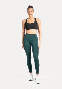 Zwarte sportbeha met brede bandjes gecombineerd met high-waisted, donkergroene leggings. Model draagt witte sportschoenen. Neutrale achtergrond.