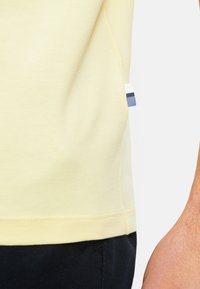 T-shirt di cotone giallo chiaro con maniche corte, con etichetta laterale a righe blu e bianche. Dettaglio di cucitura all'orlo inferiore.