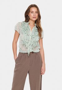 Ljussgrön skjorta med volang fram, knäppning och korta ärmar, kombinerat med bruna högmidjade byxor. Strukturerad och blommig design.