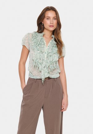 Light green blouse with ruffled front, button closure, and short sleeves, paired with brown high-waisted trousers. Textured and floral design.