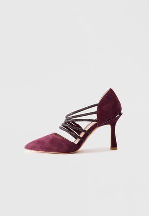Scarpa con tacco alto in suede viola, con punta affilata e numerose sottili cinghie glitterate sul lato superiore, posizionata su uno sfondo bianco semplice.
