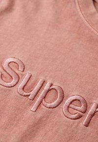 Camiseta de algodón rosa con el logotipo bordado "Super" en un tono ligeramente más oscuro. Textura suave con escote redondo y sin patrones visibles.