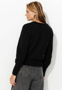 Un pull noir avec un design en tricot texturé, un ourlet et des poignets côtelés. Porté avec un jean gris taille haute, vu de dos.