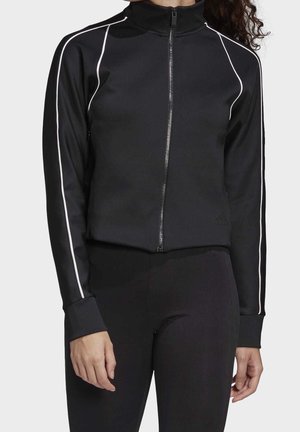 Vrouw in een zwarte jas met ritssluiting en witte bies langs de mouwen, en zwarte nauwsluitende broek, staand tegen een effen lichte achtergrond.