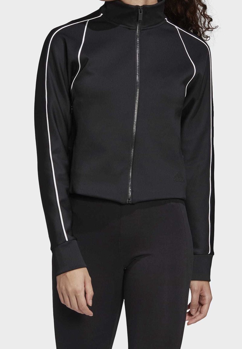 Femme portant une veste noire zippée avec un liseré blanc le long des manches et un pantalon noir ajusté, debout devant un fond clair uni.