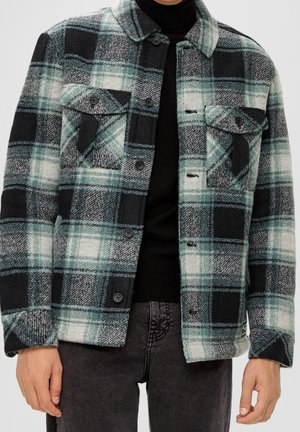 Mand iført grøn, sort og hvid ternet skjortejakke over en sort rullekrave og mørkegrå jeans, hænder afslappet langs siden.