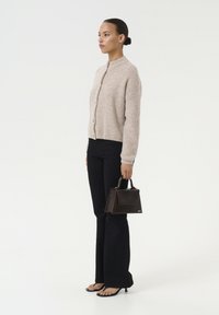 Beige, getextureerde gebreide cardigan met een hoge kraag, gecombineerd met donkere flair-jeans en zwarte haksandalen. Houdt een kleine donkere handtas vast.