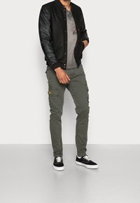 Chaqueta bomber negra con mangas de cuero, camiseta gráfica gris, pantalones cargo verde oliva con bolsillos, y zapatillas negras con cordones blancos.