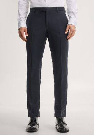 Uomo che indossa pantaloni eleganti scuri slim fit, scarpe eleganti nere lucide e una camicia bianca, in piedi davanti a uno sfondo semplice.