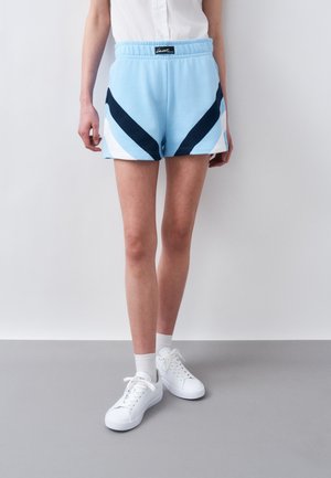 Žena v svetlomodrých športových šortkách s námorníckymi a bielymi chevrónovými pruhmi, bielych teniskách a bielej krátkej tričku s krátkym rukávom.