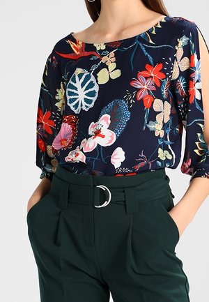 Kvinde iført en navybluse med farverigt blomsterprint og mørkegrønne, højtaljede bukser med sølvspænde, hænder i lommerne.