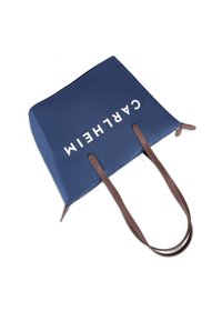 Sac fourre-tout bleu marine avec des anses en cuir marron, arborant un texte blanc audacieux "CARLHEIM." Matière lisse et brillante avec fermeture éclair en haut.