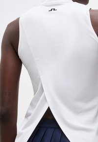 Πίσω όψη ατόμου που φοράει λευκή μπλούζα χωρίς μανίκια με διασταυρούμενη ανοιχτή πλάτη και μπλε φούστα με πιέτες.