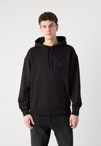 Schwarzer Kapuzenpulli mit einer Fronttasche, ausgestattet mit einem kleinen blauen Logo auf der Brust. Weicher Stoff und lässige Passform, kombiniert mit dunklen Jeans.