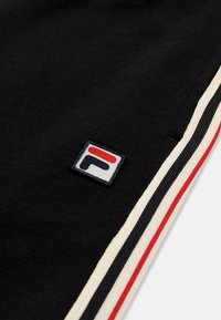 Tissu noir avec petit patch logo Fila et bandes latérales en beige, bleu marine et rouge sur une manche de vêtement ou une jambe de pantalon.