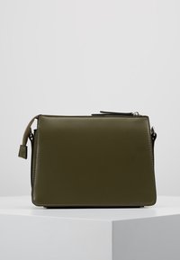 Bolso de mano rectangular de cuero verde oliva con cierre de cremallera y detalle de correa, colocado sobre un pedestal blanco contra un fondo claro sencillo.