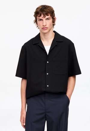 Giovane uomo con capelli castani ondulati indossa una camicia nera a maniche corte abbottonata sopra una maglietta bianca e pantaloni scuri, in piedi con una mano in tasca.