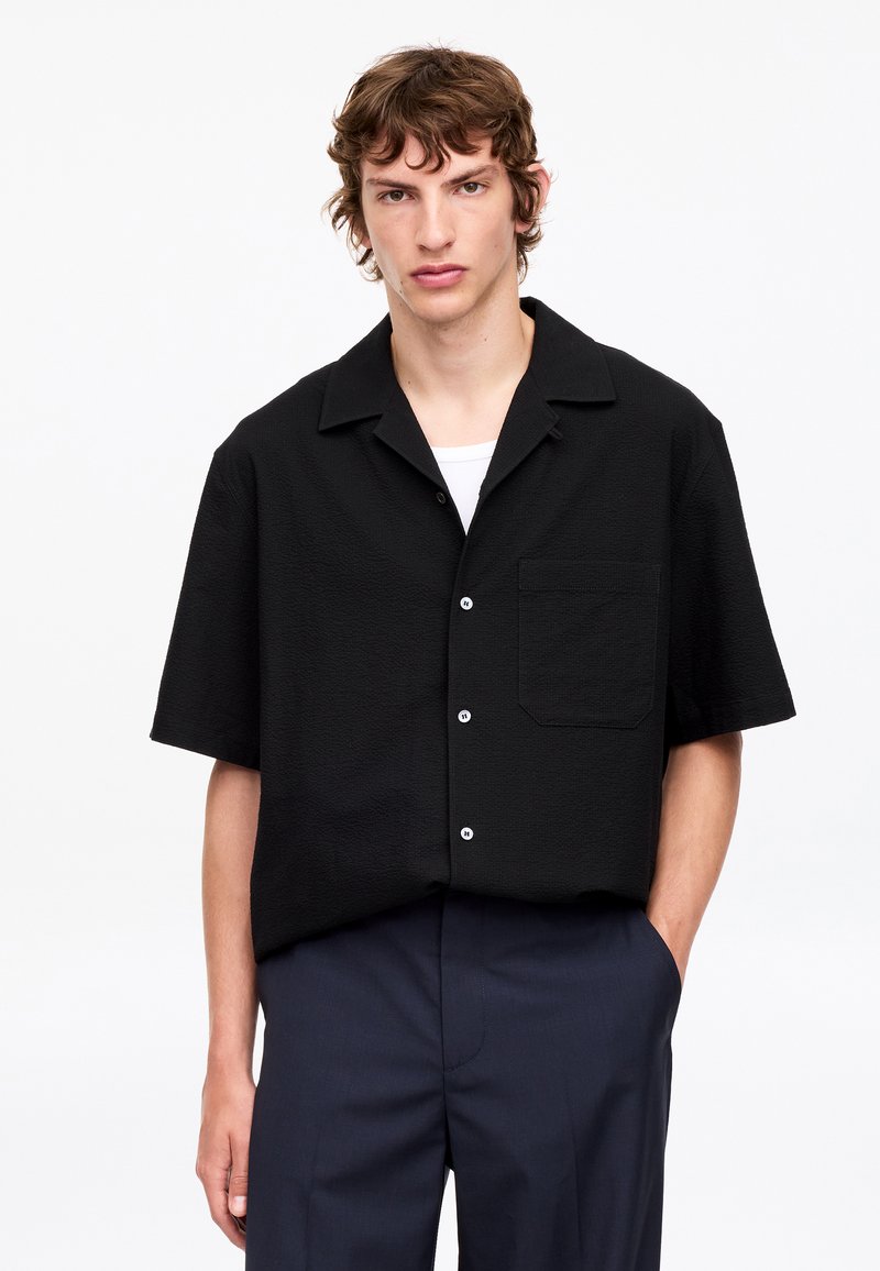 Jeune homme aux cheveux bruns ondulés porte une chemise noire à manches courtes boutonnée sur un haut blanc et un pantalon foncé, debout avec une main dans la poche.