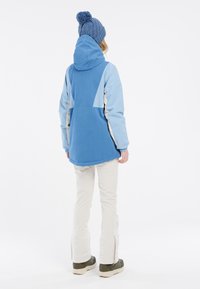 Giacca outdoor blu con cappuccio, maniche blu più chiare, dettagli beige e una vestibilità rilassata, abbinata a pantaloni da neve crema e stivali scuri.