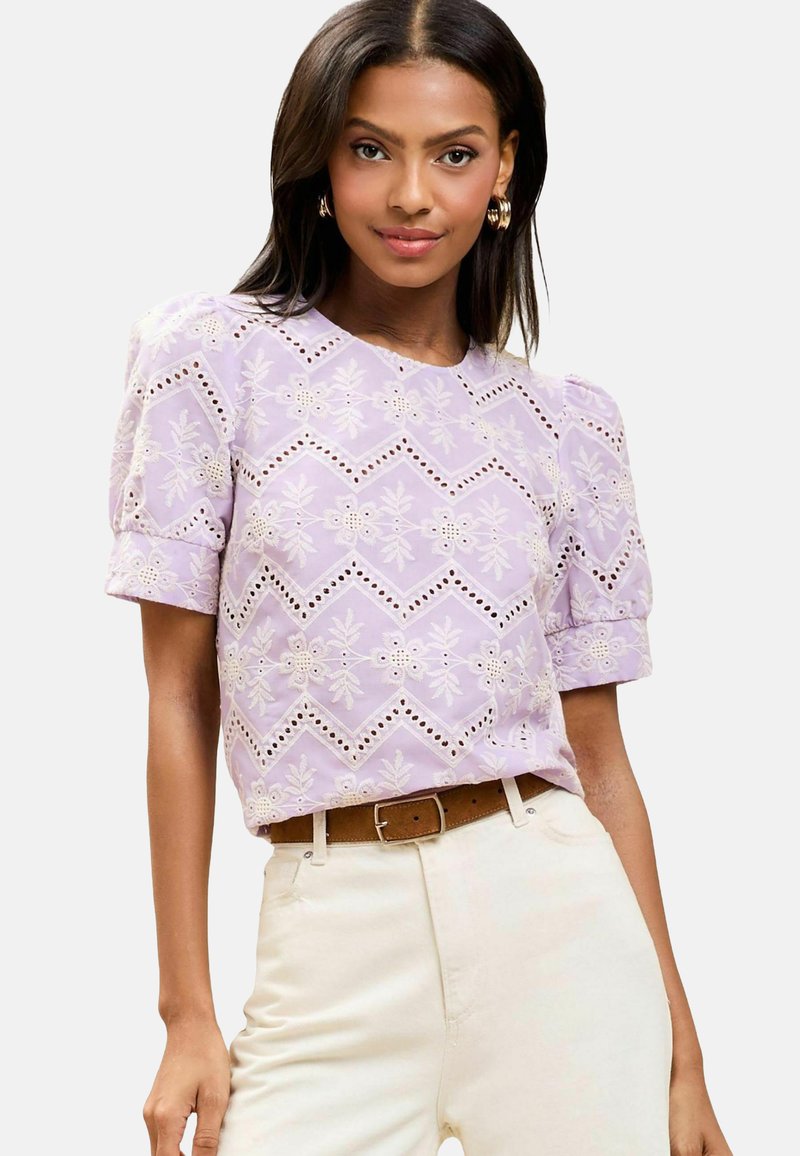 Lila Bluse mit kurzen Ärmeln, die mit gestickten Blumenmustern und Lochdetails verziert ist und einen Rundhalsausschnitt hat. Kombiniert mit beigen, hochtaillierten Hosen.