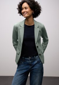 Blazer vert clair texturé à coupe ajustée, associé à une chemise bleu marine foncé et un jean bleu à coupe décontractée. Dispose de deux poches avant.