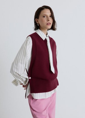 Giovane donna con capelli corti e castani indossa una camicia bianca a righe, un maglione senza maniche color bordeaux, pantaloni rosa e orecchini a cerchio dorati su sfondo uniforme.