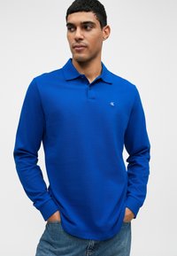 Modrá polokošeľa s dlhým rukávom vyrobená z textúrovaného bavlneného zmesi, s klasickým golierom a malým logom na ľavej strane hrude, skombinovaná so svetlým denimu.