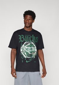 Sort t-shirt med "Milwaukee Bucks" i fede grønne bogstaver, forsynet med et grafisk motiv af en basketball og Bucks-logoet i hvide og grønne detaljer.
